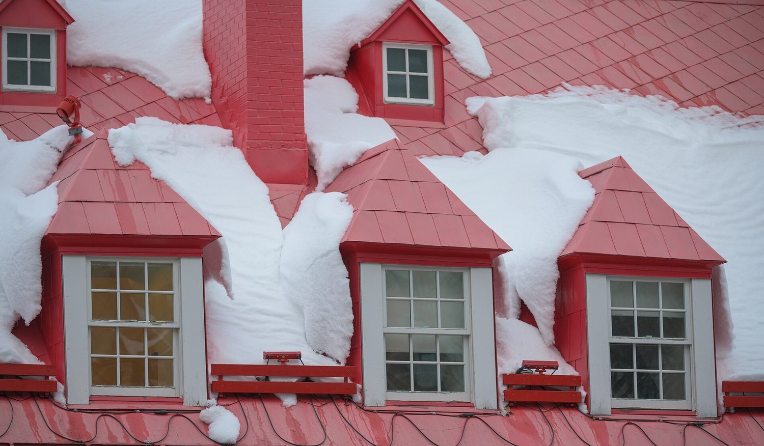 A red metal roof with dormers where snow has partly slid off in sheets, with snow retention bars fitted below the dormers